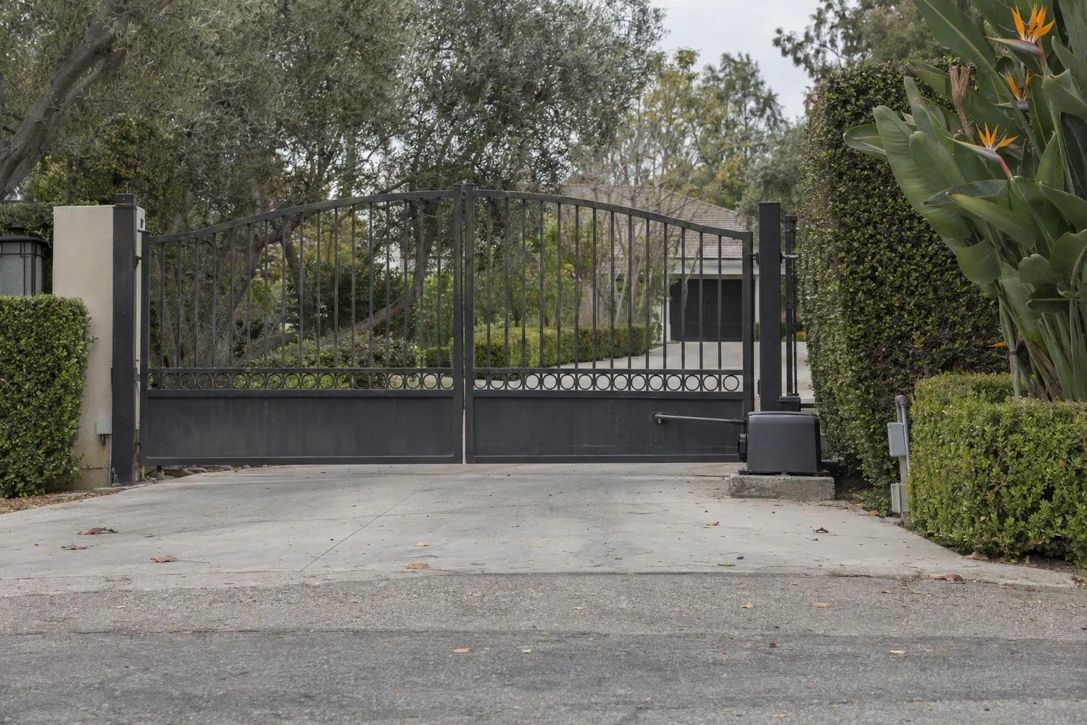 A gate opener motor mounted on a concrete pad beside a closed wrought iron driveway gate, with a gravel driveway and trimmed shrubs around a Southern California home in daylight