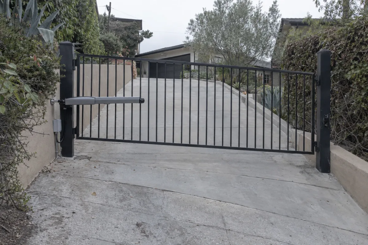 A black wrought iron swing gate on a steep residential driveway, with a gate opener arm mounted to the post and the concrete driveway sloping sharply downhill toward the street