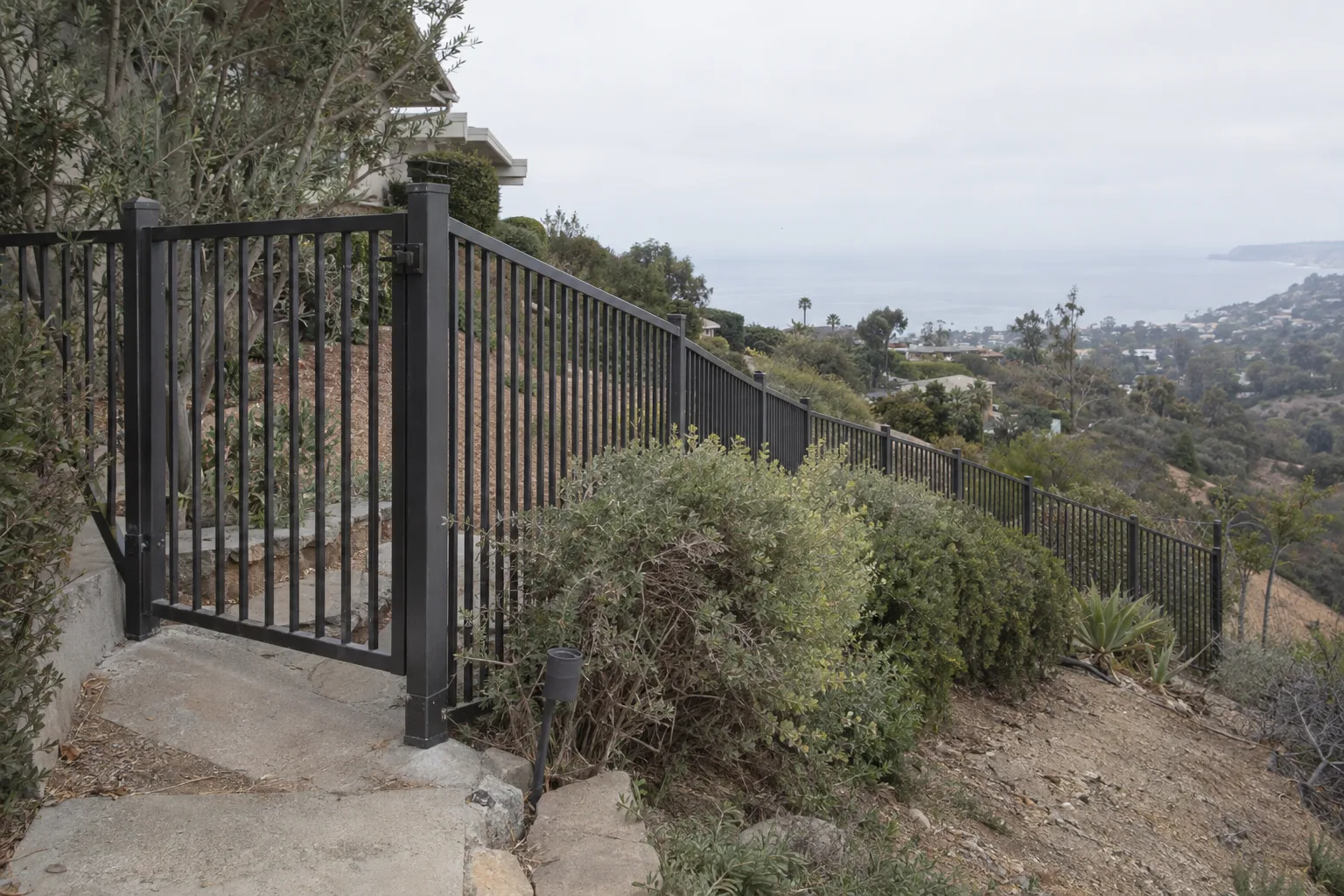 Powder-coated aluminum fence and pedestrian gate running along a hillside property line with ocean haze visible in the background and clipped landscaping beside the fence