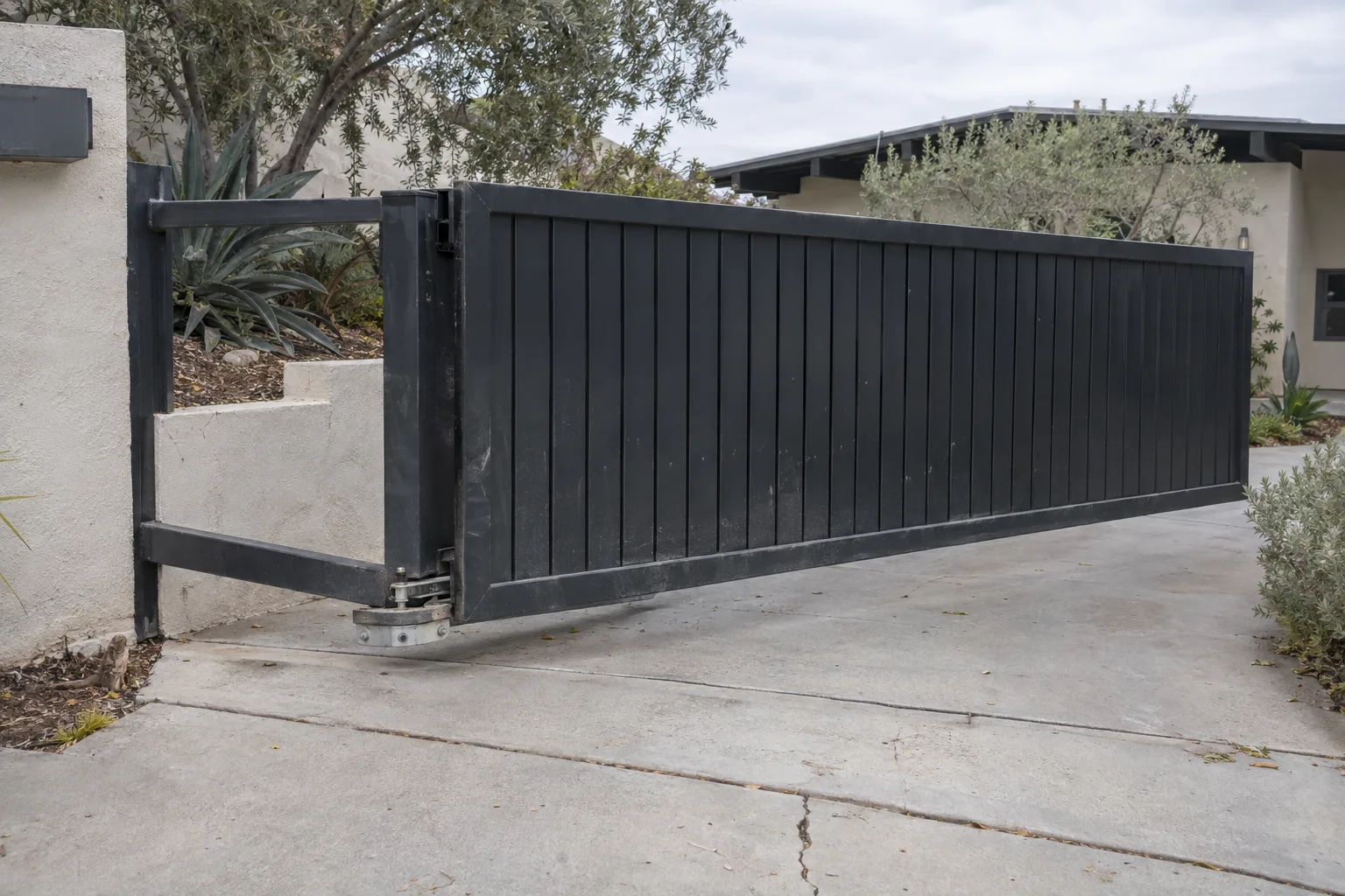 A black steel cantilever sliding gate partially open across a sloped concrete driveway, with the gate suspended above the ground and the roller carriage and counterbalance section visible at the support post beside a stucco wall.