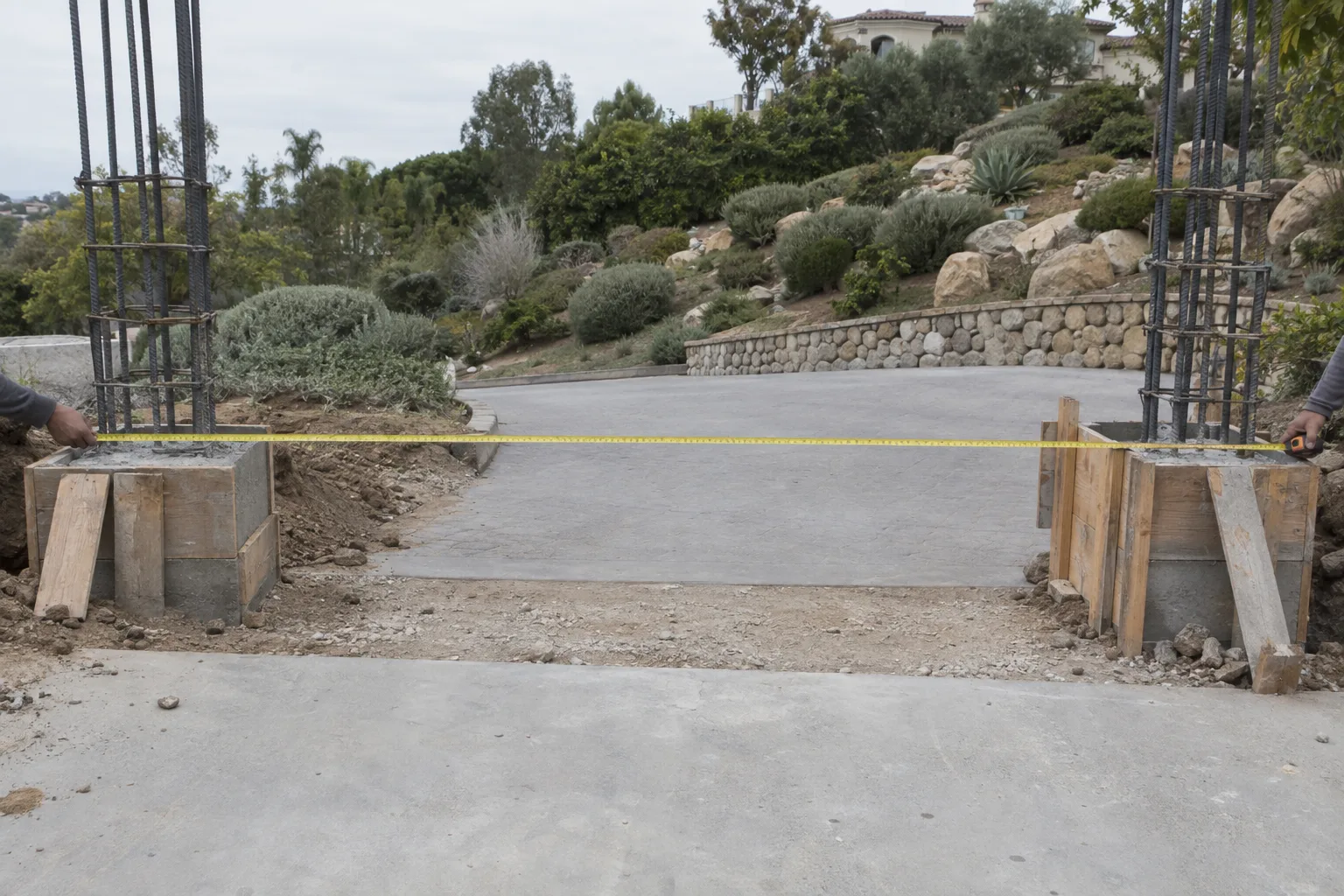 Two concrete gate pillar footings under construction at a driveway entrance, vertical rebar protruding from each formed base, a tape measure spanning the opening between them, with hillside estate landscaping in the background.
