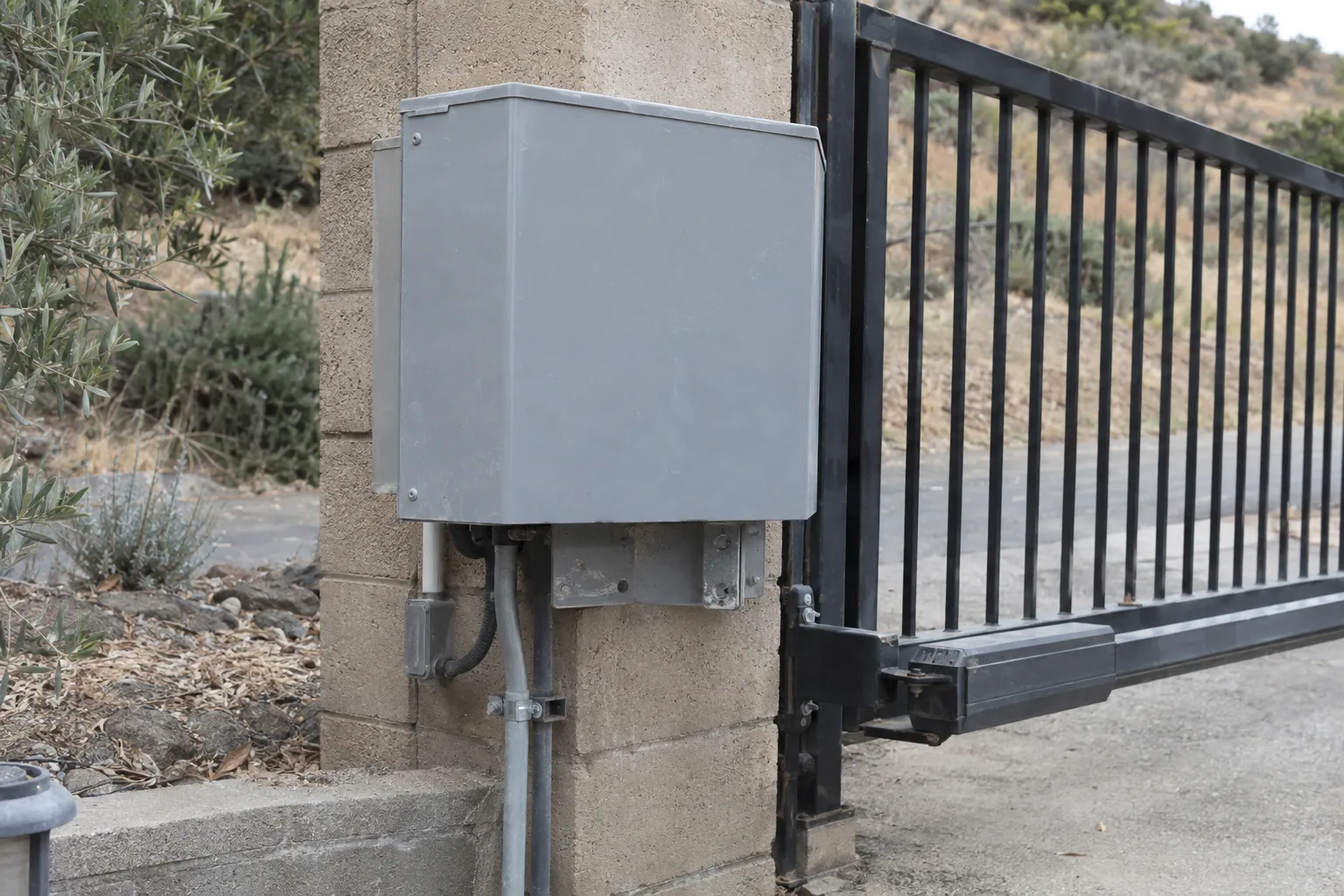Close view of a weatherproof gate operator box mounted on a block post in bright Southern California sun, with conduit wiring and a mounting bracket visible and a dry hillside landscape behind
