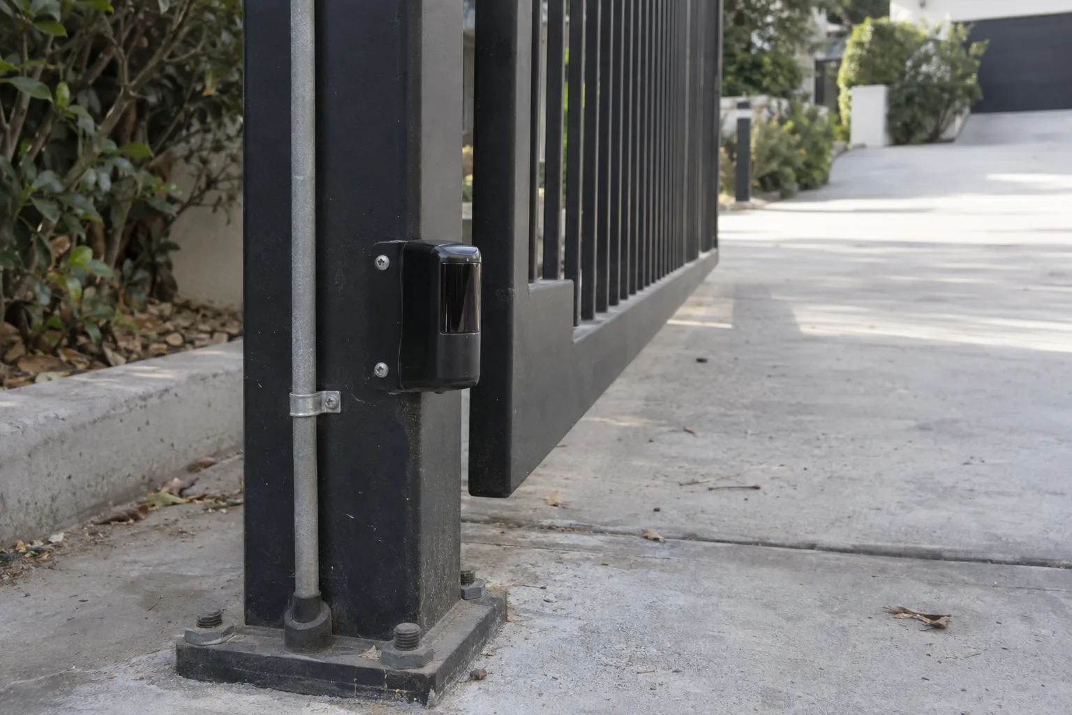 A black infrared safety beam sensor mounted near the base of a steel gate post, with wiring routed neatly down the post, and a wrought iron gate leaf and concrete driveway visible behind