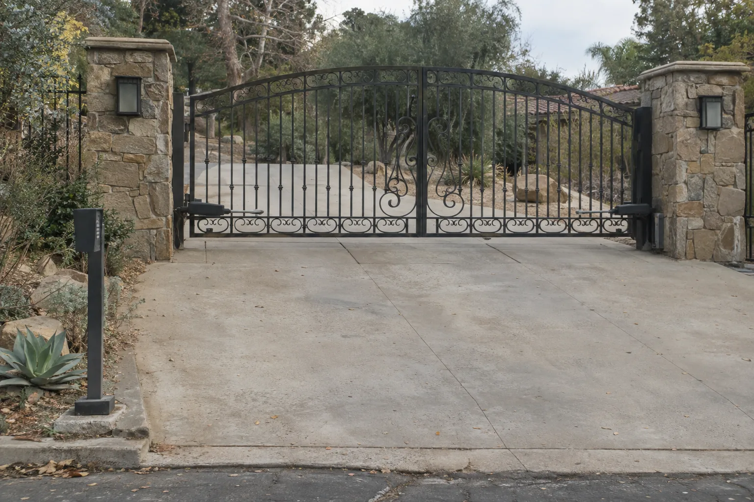 Closed wrought iron double swing driveway gate with ornamental pickets, flanked by stone pillar columns, a keypad post at the kerb, bathed in warm late-afternoon sunlight on a residential Los Angeles driveway.