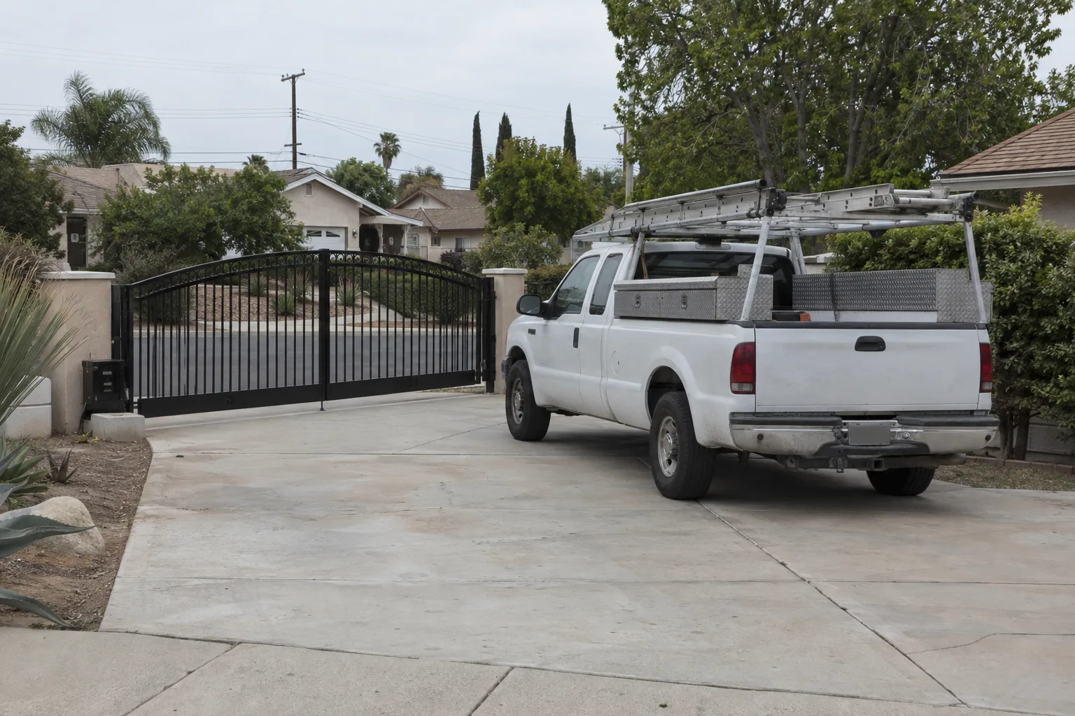 A white work truck equipped with ladder racks and tool storage bins parked next to a black wrought iron swing gate in a residential driveway, with a sunny San Fernando Valley street and stucco homes visible in the background.