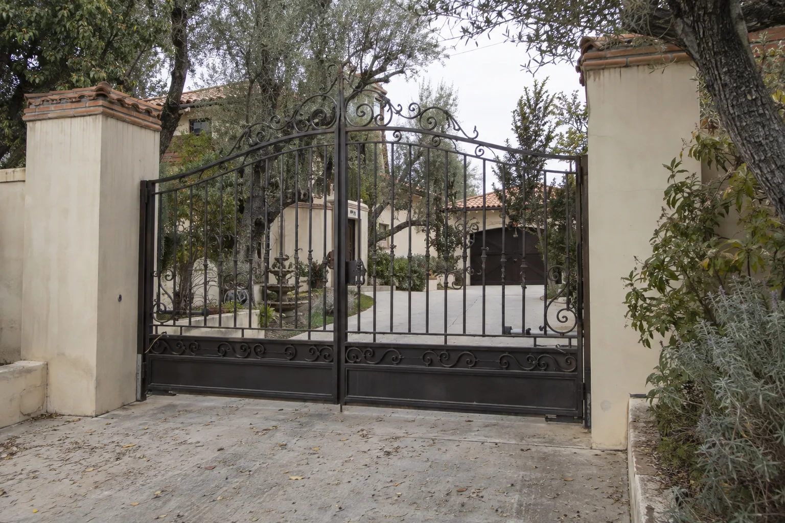 Tall arched wrought iron driveway gate with scroll detailing, flanked by cream stucco pilasters with terracotta tile caps, at a Spanish colonial home entry in afternoon sunlight