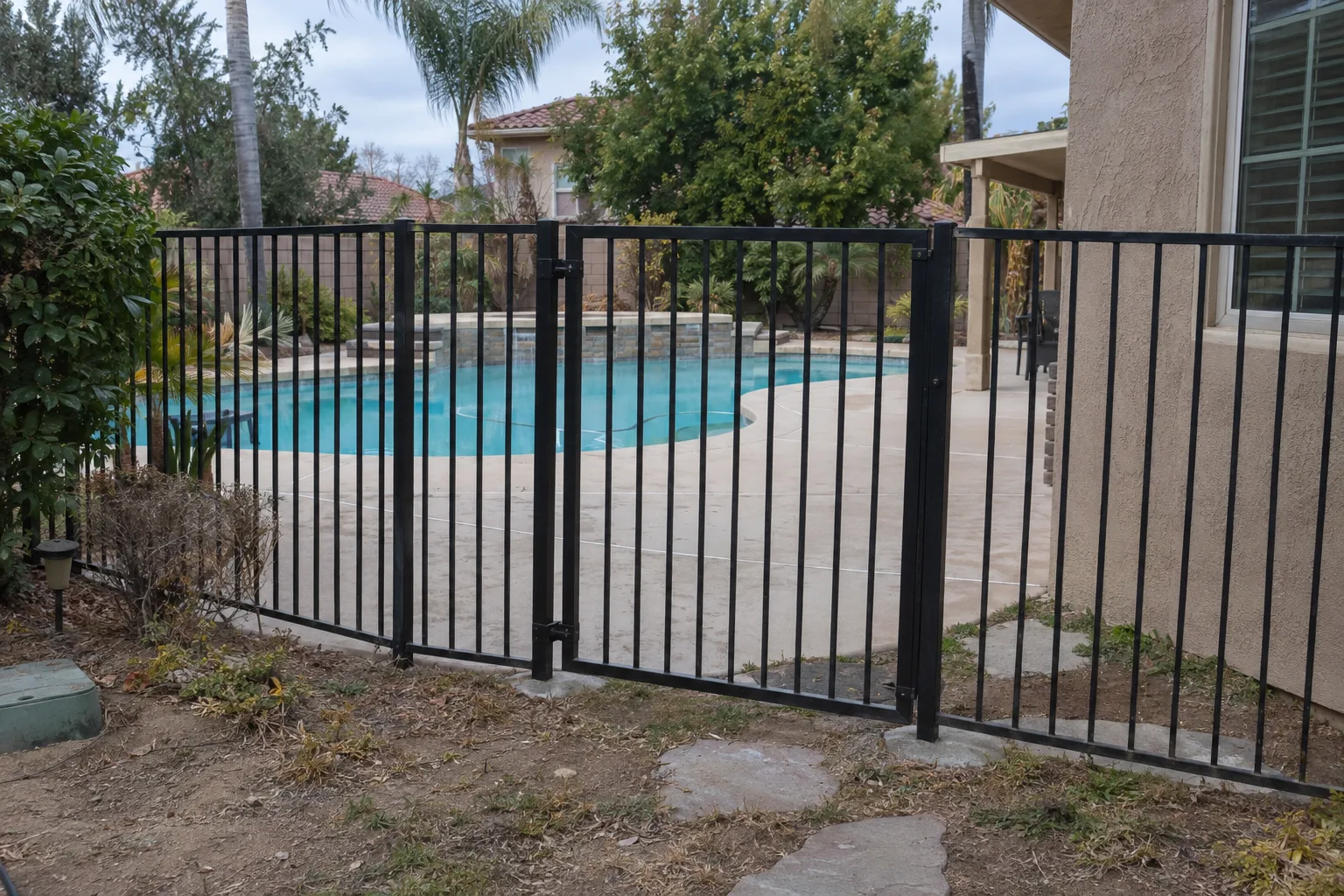 Black aluminum picket pool fence enclosing a backyard swimming pool, with a self-latching gate in the barrier and a blue pool and concrete patio visible beyond, under Southern California daylight