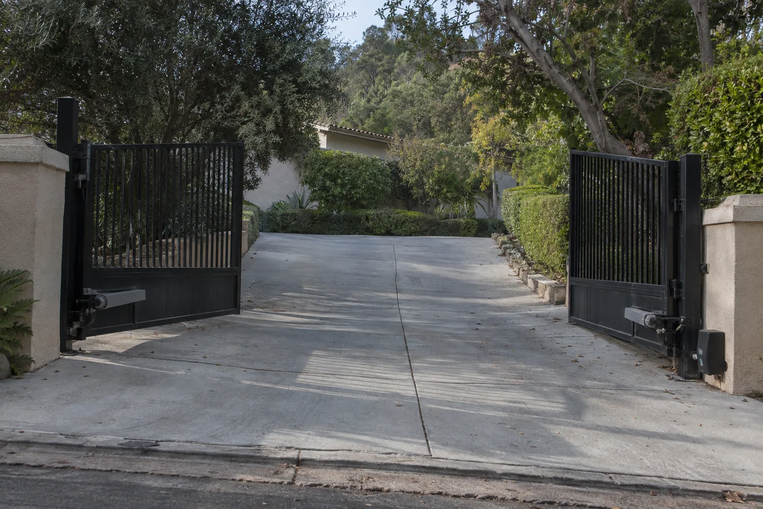 A tall black steel automatic driveway gate standing open at the entrance of a hillside home, with a sloping concrete driveway, stucco wall, and mature hedges visible in warm late-afternoon sunlight.