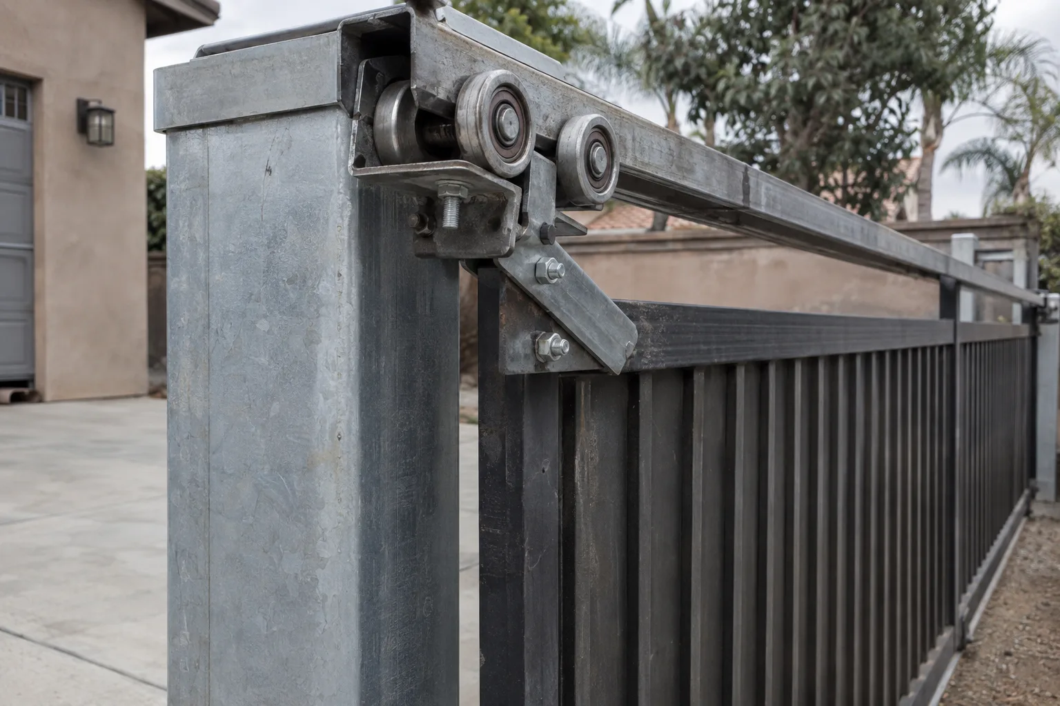 Steel cantilever sliding gate carriage and rollers clamped to an overhead rail on a galvanized post at a residential driveway, with bare concrete below and no ground track