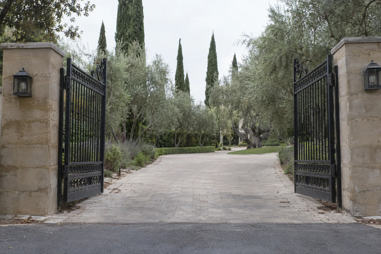A pair of open wrought iron swing gate leaves flanked by stone pillars at a wide estate driveway, mature trees lining the property behind them in afternoon sunlight