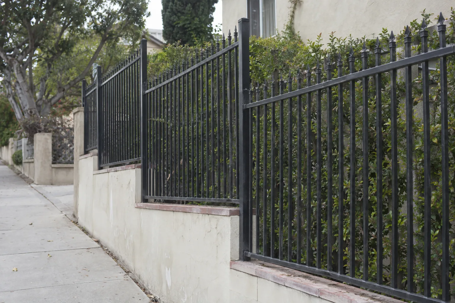 Black tubular steel ornamental fence with spear-top finials following the slope of a residential property line, with a trimmed hedge and cream stucco wall behind it under Los Angeles daylight