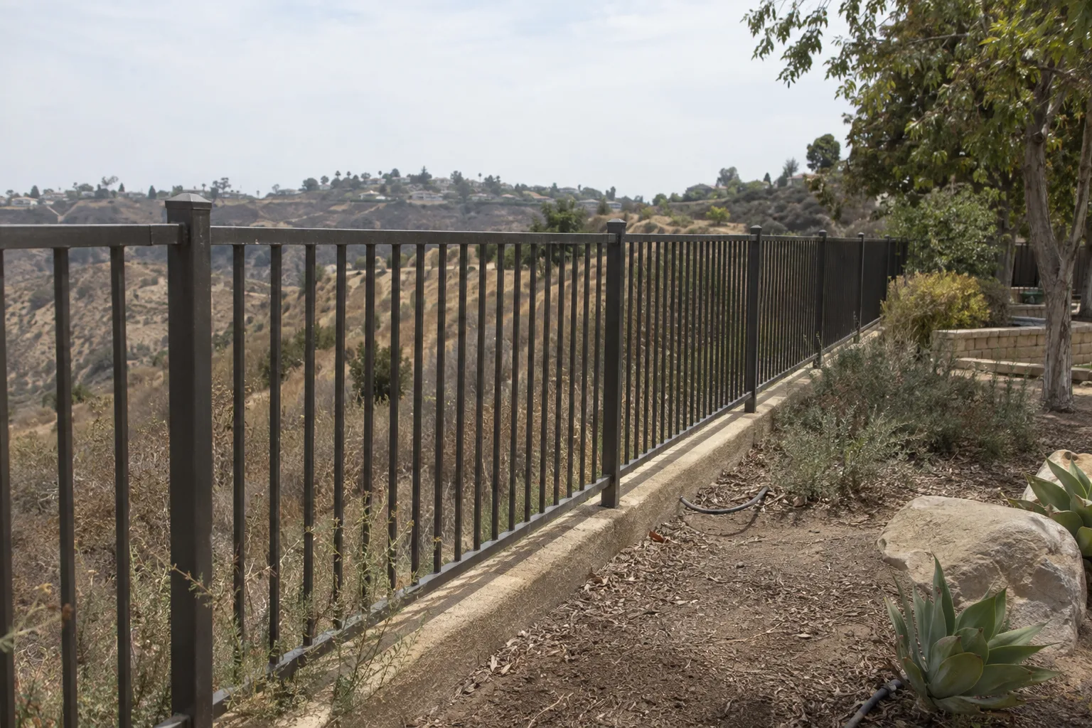 A wrought iron fence running along the edge of a hillside lot in the Southern California foothills, with dry chaparral brush and scrub vegetation visible just beyond the fence line on the sloping terrain behind it