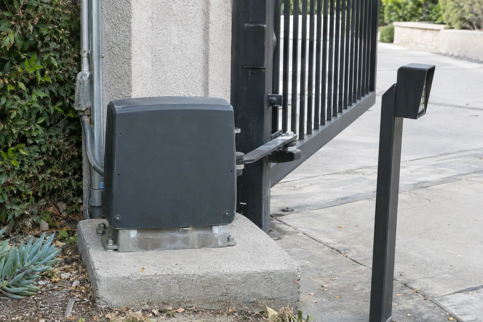 A gate operator motor unit mounted on a concrete pad next to a driveway gate post, with grey conduit running along the post and a keypad pedestal standing nearby on the driveway apron