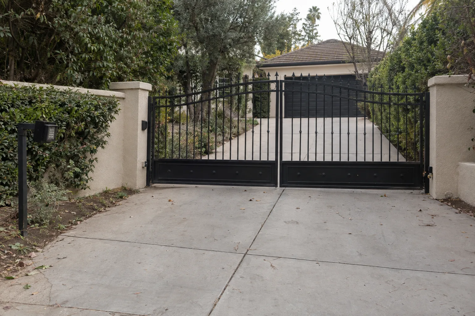 A closed black wrought iron driveway gate spanning a residential entrance in Encino, with a keypad post at the kerb, stucco walls and a hedge on either side, lit by warm late-afternoon sunlight
