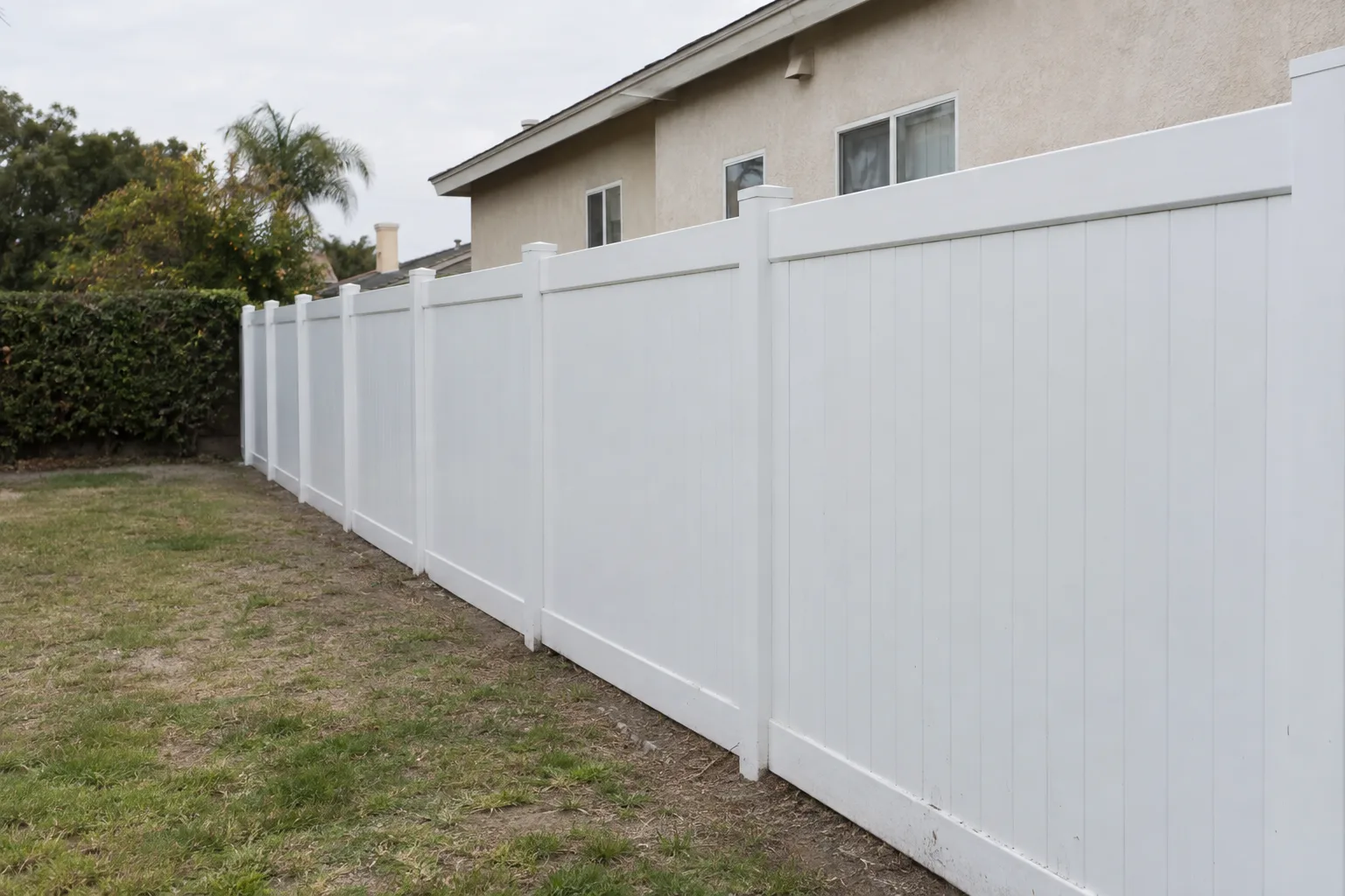 A white vinyl privacy fence running along a suburban backyard property line in bright daylight, with a stucco house and trimmed hedge visible in the background.