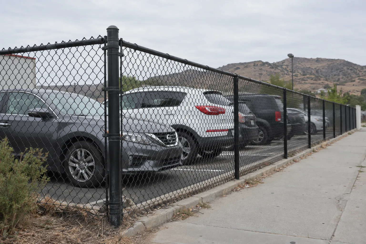 Black vinyl-coated chain link fence along a commercial parking lot perimeter in Southern California, with a terminal corner post, top rail, and several parked cars visible behind the mesh under midday sun