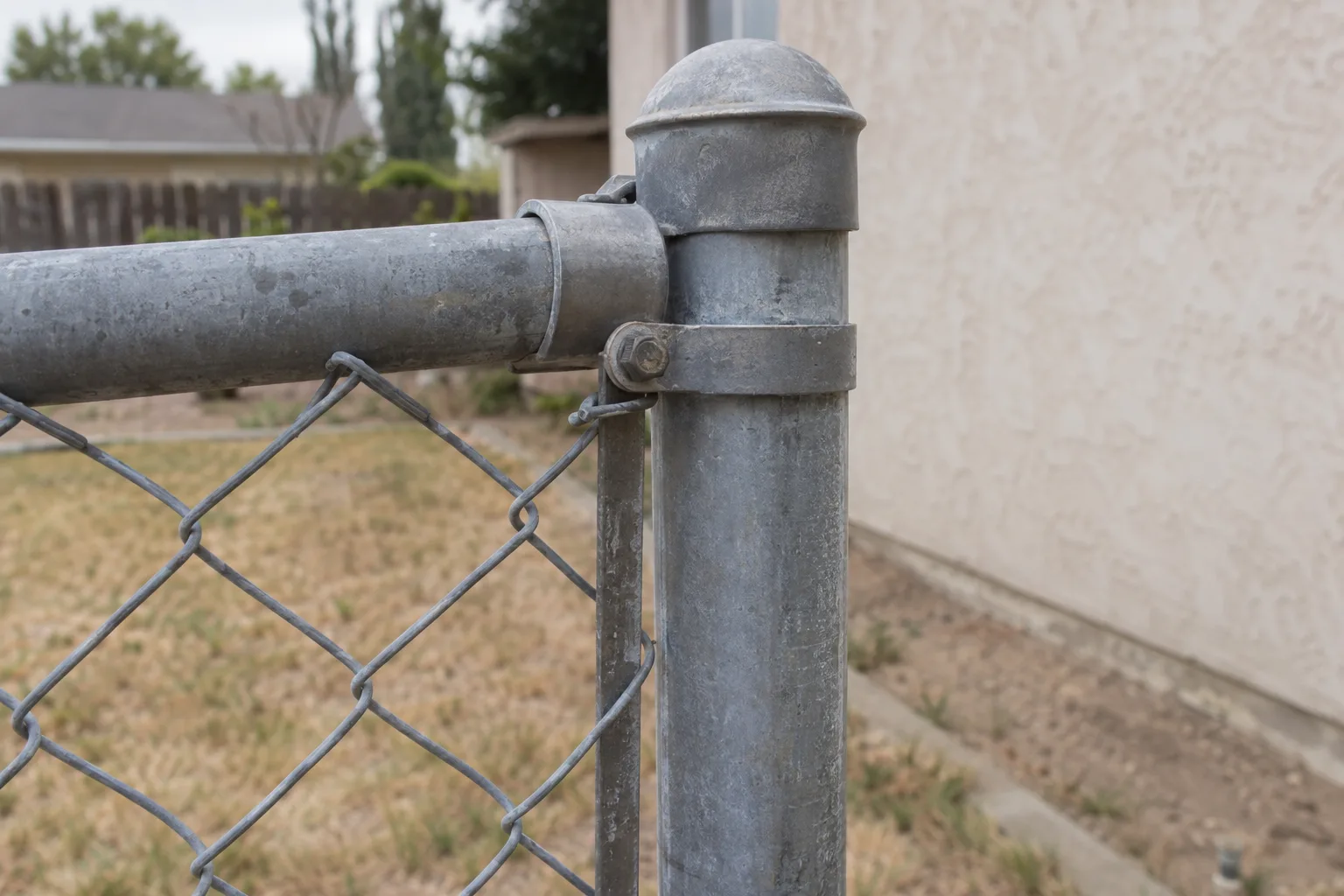 Close-up of a galvanized chain link fence terminal post with a tension band clamp and top rail fitting, diamond mesh fabric stretched taut, set against a blurred Southern California residential backyard