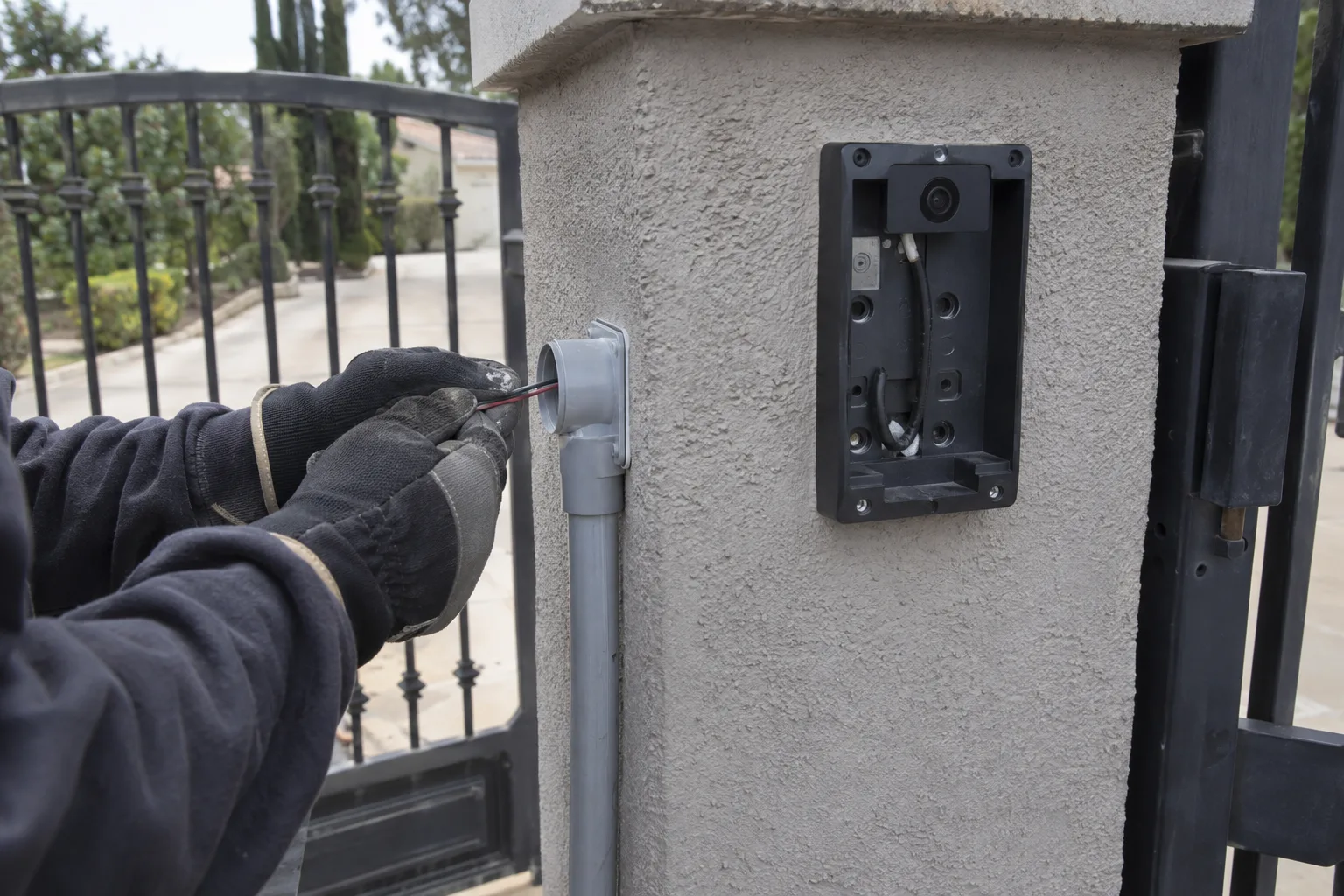 Gloved hands routing low-voltage wiring through a conduit connector on the side of a stucco gate post, with a video intercom backplate mounted on the post face and a wrought iron driveway gate blurred in the background.