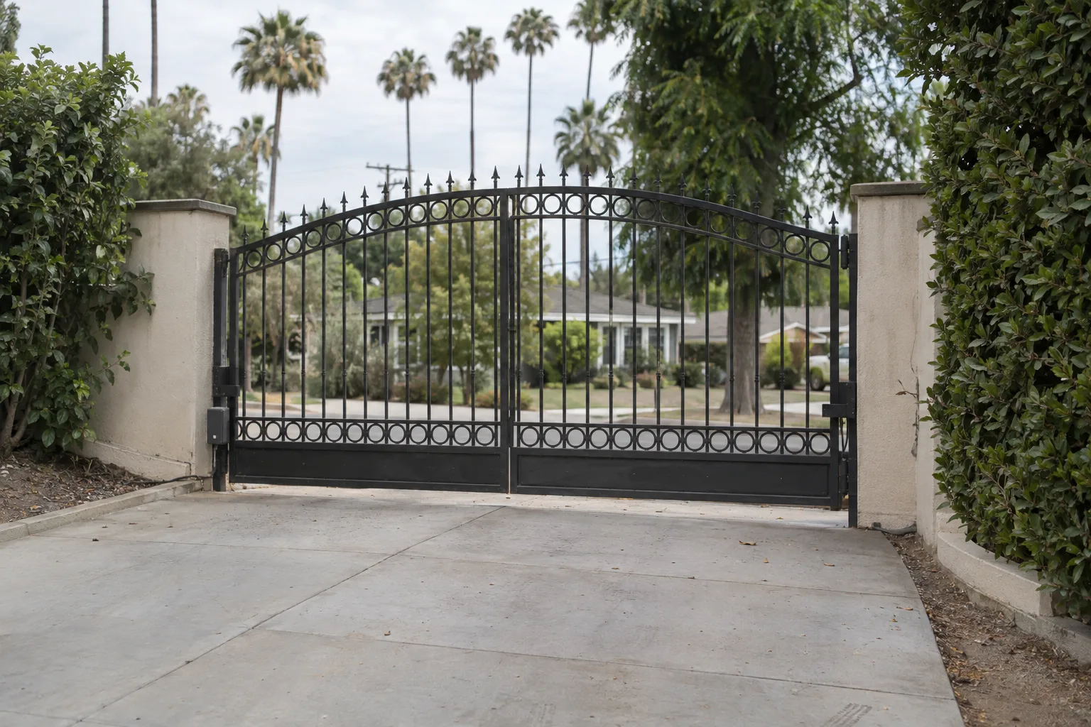 A closed dark wrought iron driveway gate spanning a residential driveway, with a stucco perimeter wall and trimmed hedge on each side, photographed in bright afternoon sunlight on a wide San Fernando Valley street