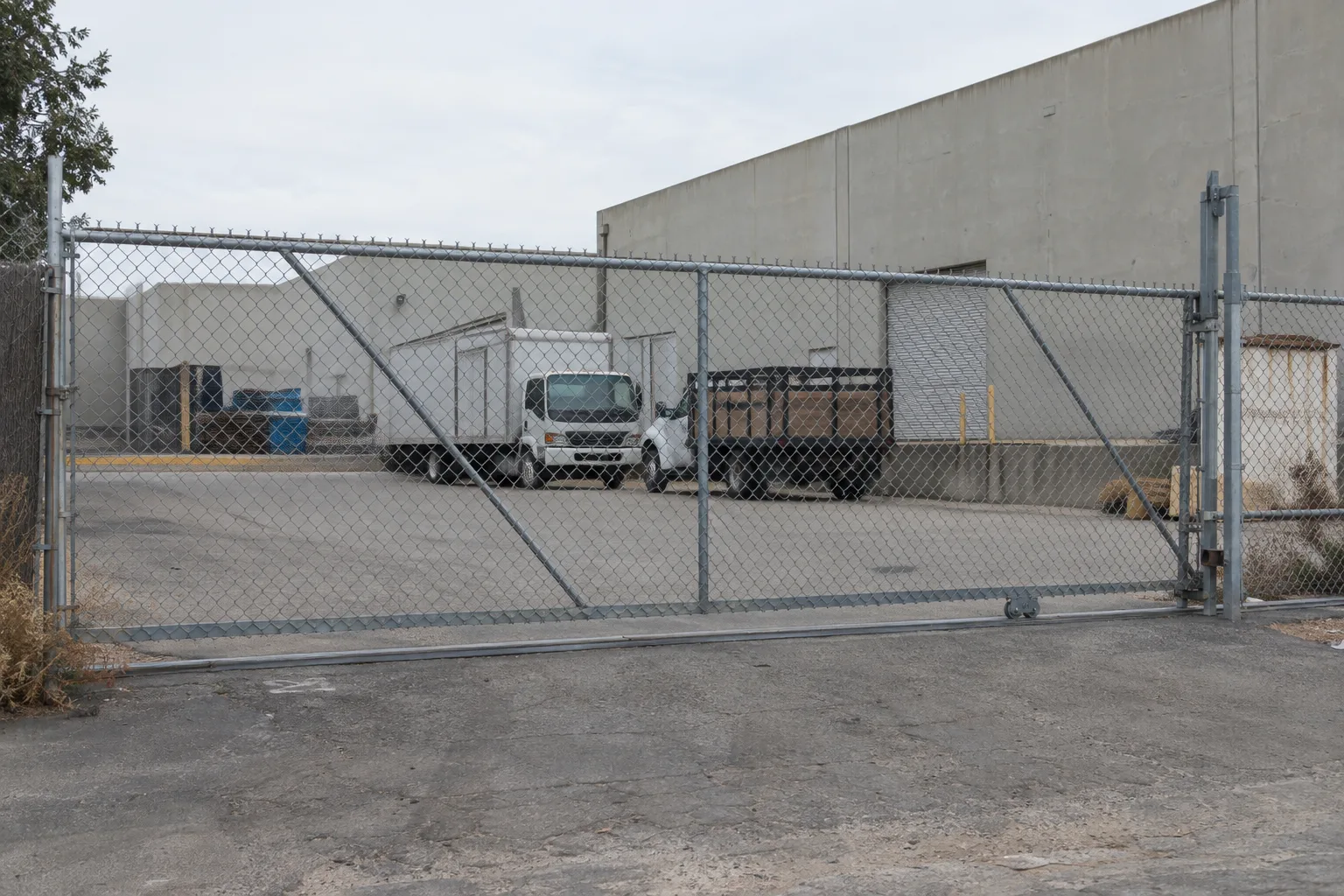 A closed heavy-gauge chain link sliding gate at a commercial warehouse yard entrance, with box trucks parked near a loading dock and tilt-up concrete industrial buildings visible behind the fence in a Southern California industrial setting.
