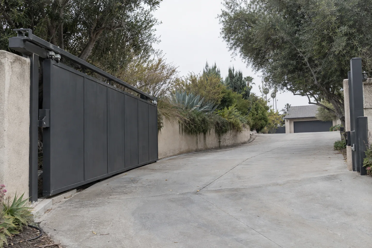 A dark cantilever sliding gate in the open position alongside a sloped residential driveway, showing the overhead horizontal rail and carriage hardware mounted to a steel post with no ground track, framed by a stucco wall and drought-tolerant plantings under bright Southern California sunlight.