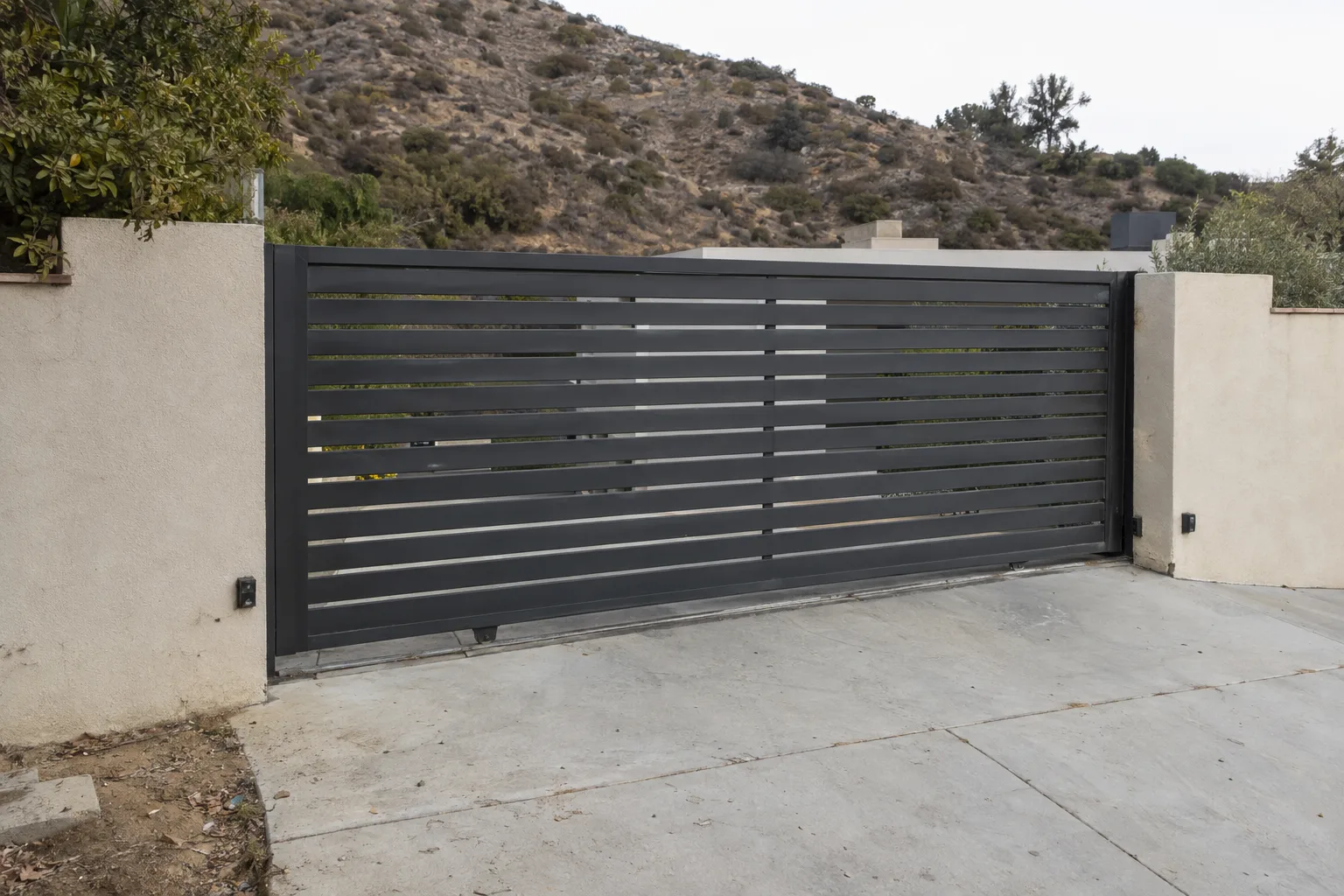 A closed dark steel sliding driveway gate spanning a sloped residential driveway, with a visible grade change beneath the gate panel, a stucco wall on one side, and a dry chaparral hillside in the background under bright afternoon sun.