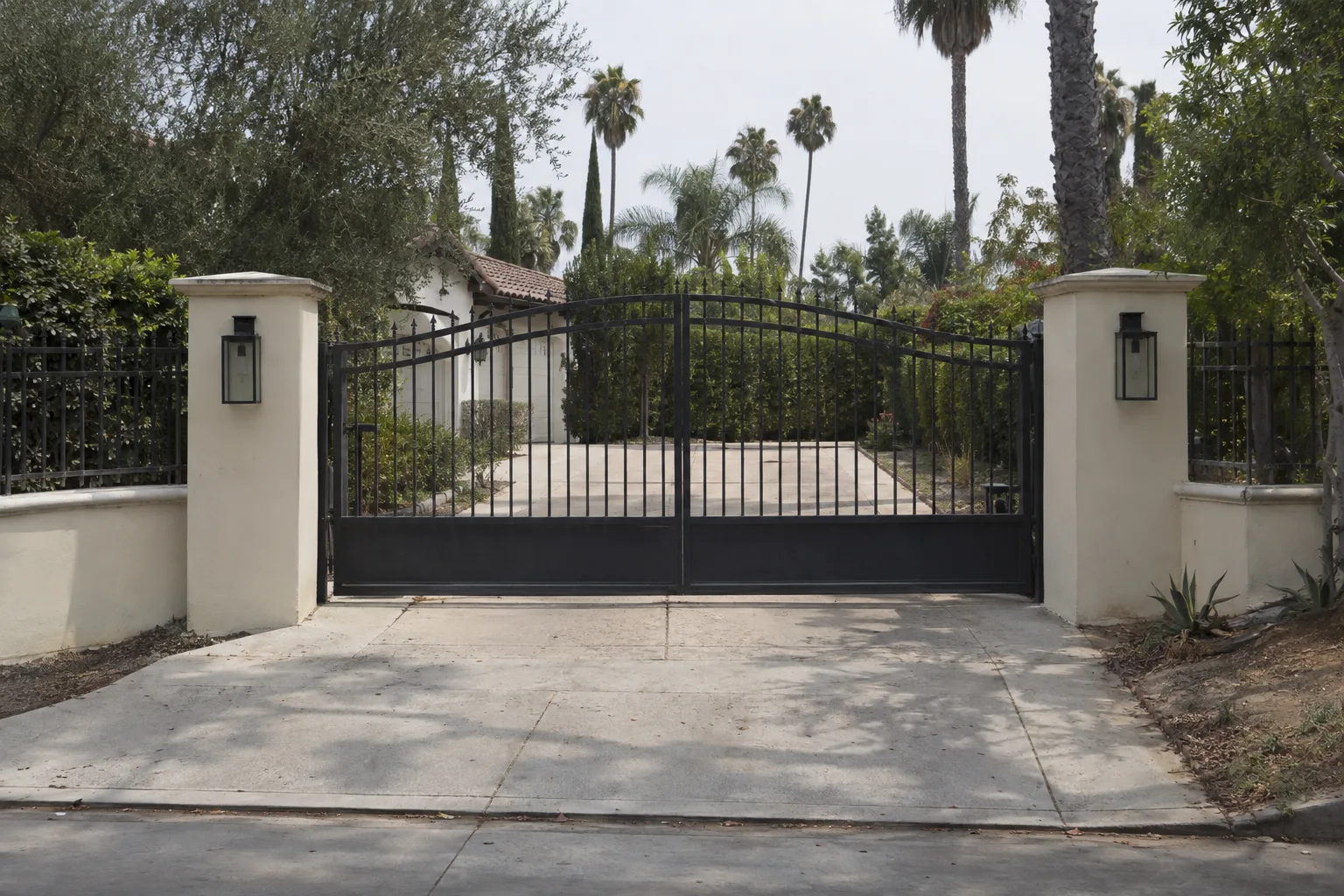 Closed dark wrought iron driveway gate with matching iron fence panels attached to stucco pillars along a low stucco perimeter wall of a residential property on a sunny Southern California street
