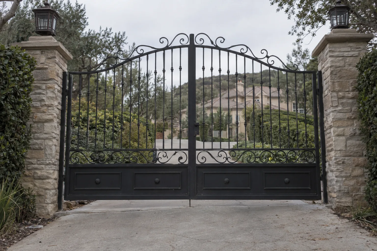 Tall closed wrought iron double-leaf estate gate with ornamental vertical pickets flanked by stone pillars, manicured green hedges on each side, a long driveway leading to a hillside home visible in the background under warm late-afternoon light.