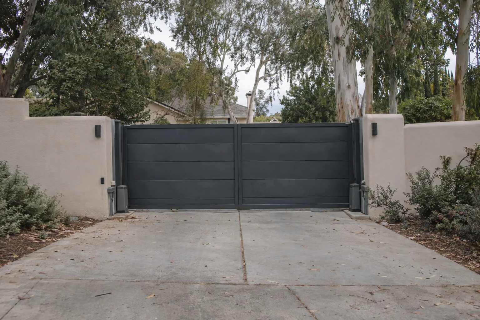 Wide view of a closed dark steel automatic driveway gate across a residential entrance, with a concrete driveway leading up to it, smooth stucco walls on either side, and mature trees under clear Southern California daylight.