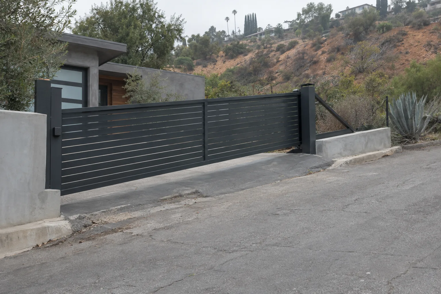A dark steel cantilever sliding gate mounted on posts beside a steeply sloped residential driveway, suspended above the inclined asphalt with no ground track, a low concrete retaining wall and dry hillside terrain visible in the background under bright Los Angeles midday light.