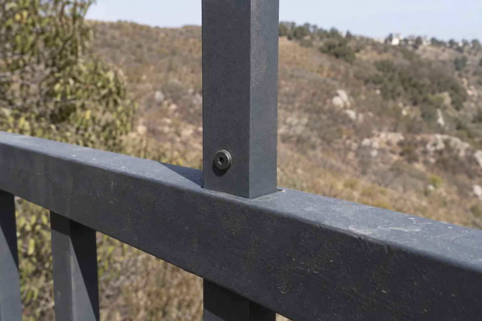 Close-up of a dark charcoal aluminum fence picket connected to a horizontal rail in bright sunlight, showing the matte powder-coated metal texture and fastener detail, with a dry hillside blurred in the background